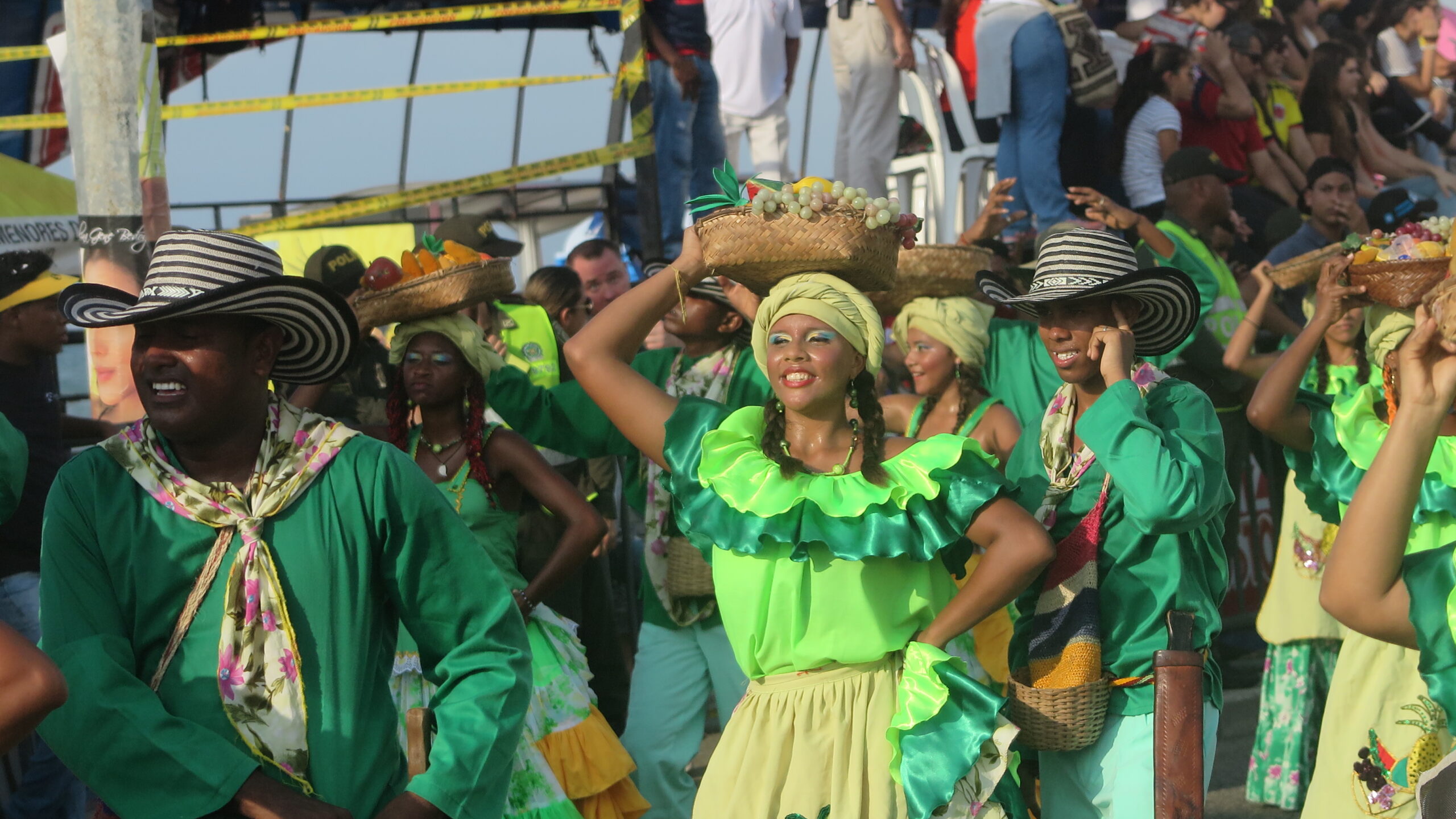 Participants in a parade in Cartagena, Colombia, wearing traditional green and yellow costumes. Women balance baskets of fruit on their heads while men wear "sombrero vueltiao" hats.