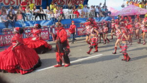 A parade in Cartagena, Colombia, featuring dancers in elaborate red and black costumes. Women wear large, shimmering red dresses, while a man in a red suit and mask walks with a staff.