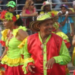 Smiling dancers in vibrant red, lime green, and yellow traditional costumes during a parade in Cartagena. A man in a red and green satin shirt and a traditional hat stands in the center.