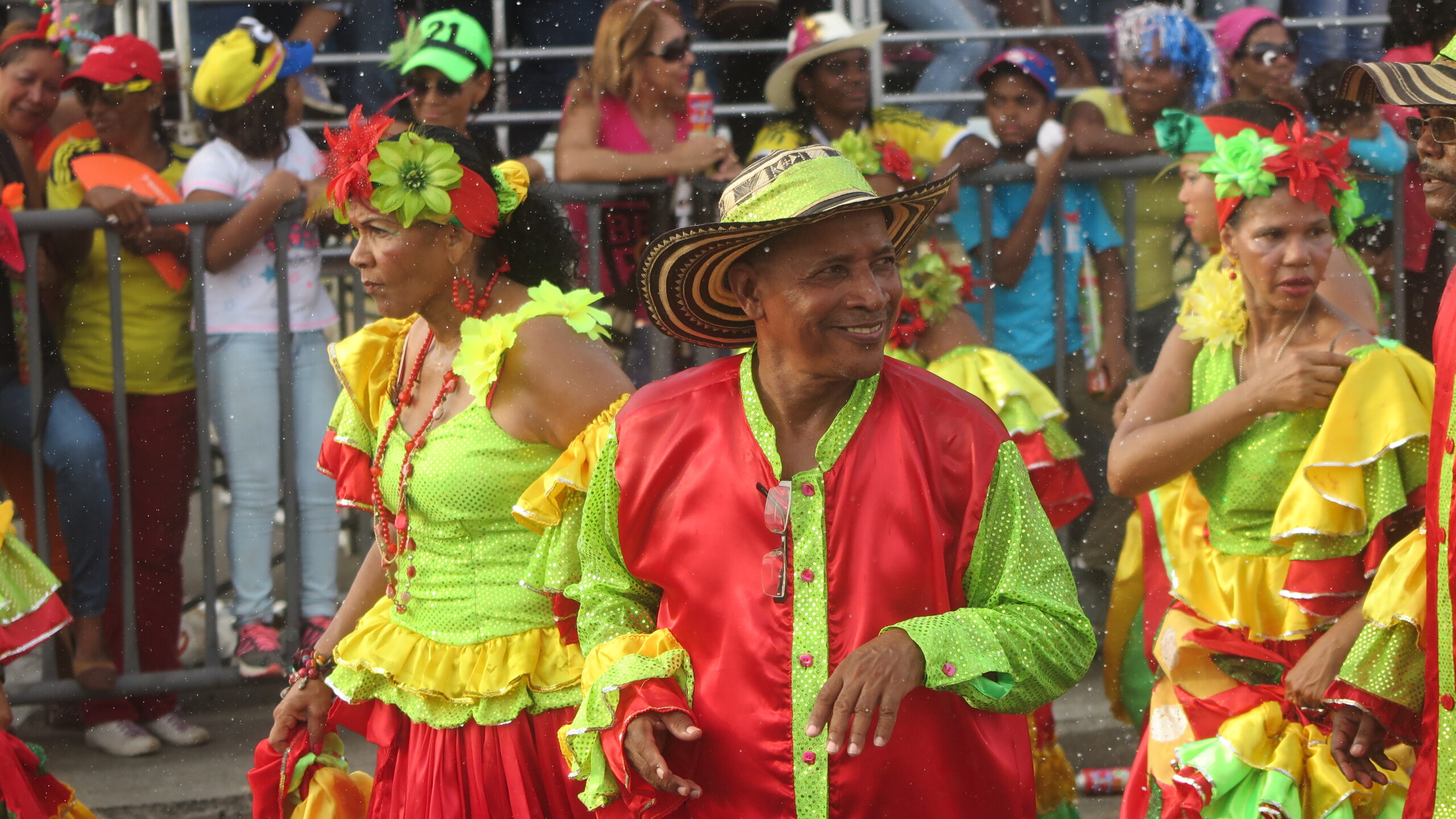 Smiling dancers in vibrant red, lime green, and yellow traditional costumes during a parade in Cartagena. A man in a red and green satin shirt and a traditional hat stands in the center.