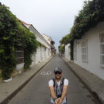 Wide-angle selfie of a man in a narrow Cartagena street with white colonial buildings and green vines.