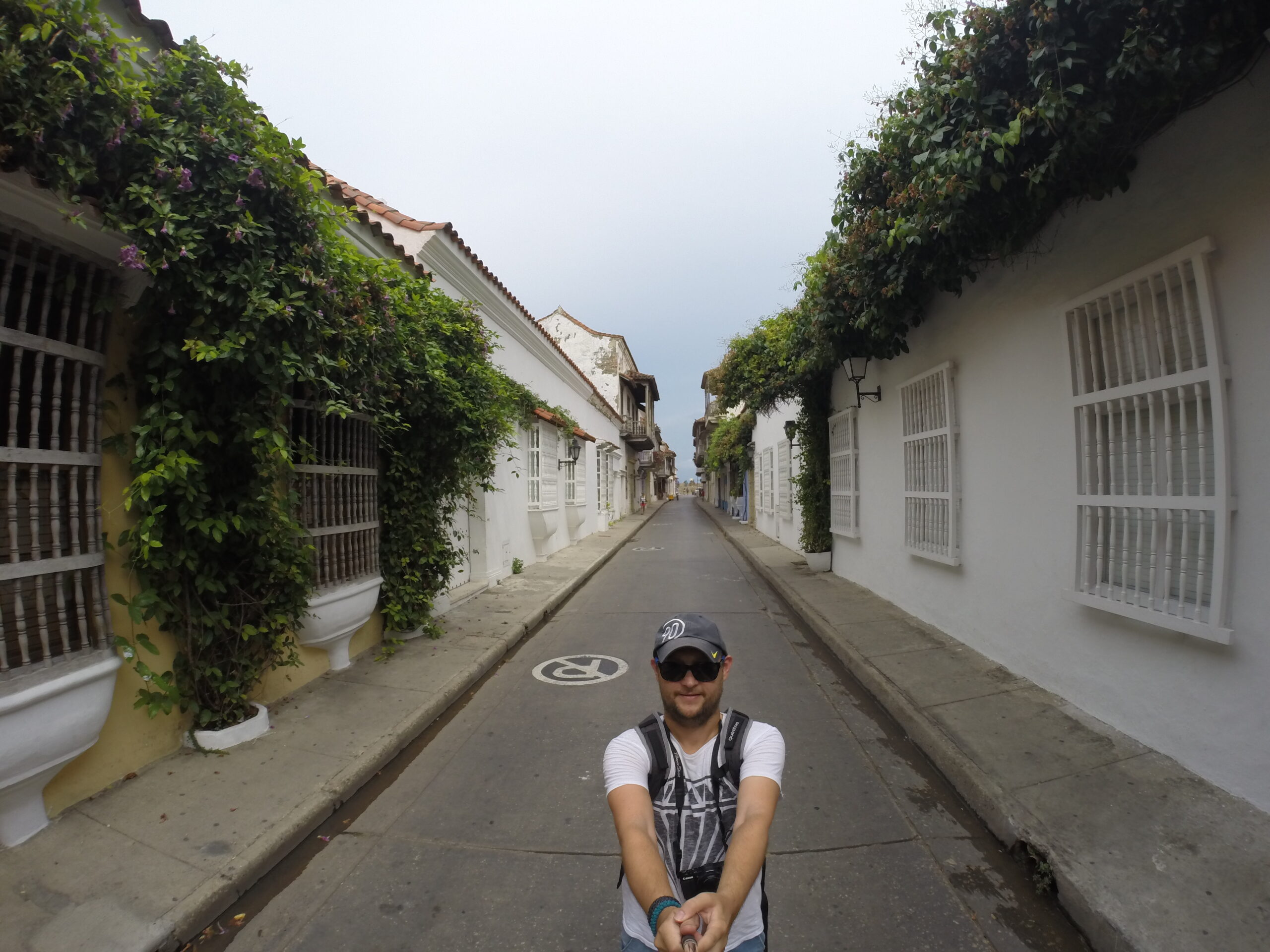 Wide-angle selfie of a man in a narrow Cartagena street with white colonial buildings and green vines.