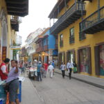 A lively street in Cartagena with colorful yellow and blue buildings, local vendors, and pedestrians.