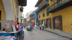 A lively street in Cartagena with colorful yellow and blue buildings, local vendors, and pedestrians.
