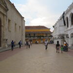 People walking through a wide, open plaza in Cartagena flanked by historic stone and white arched buildings.