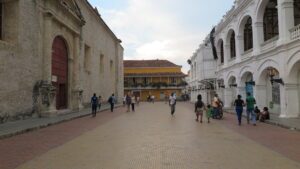 People walking through a wide, open plaza in Cartagena flanked by historic stone and white arched buildings.