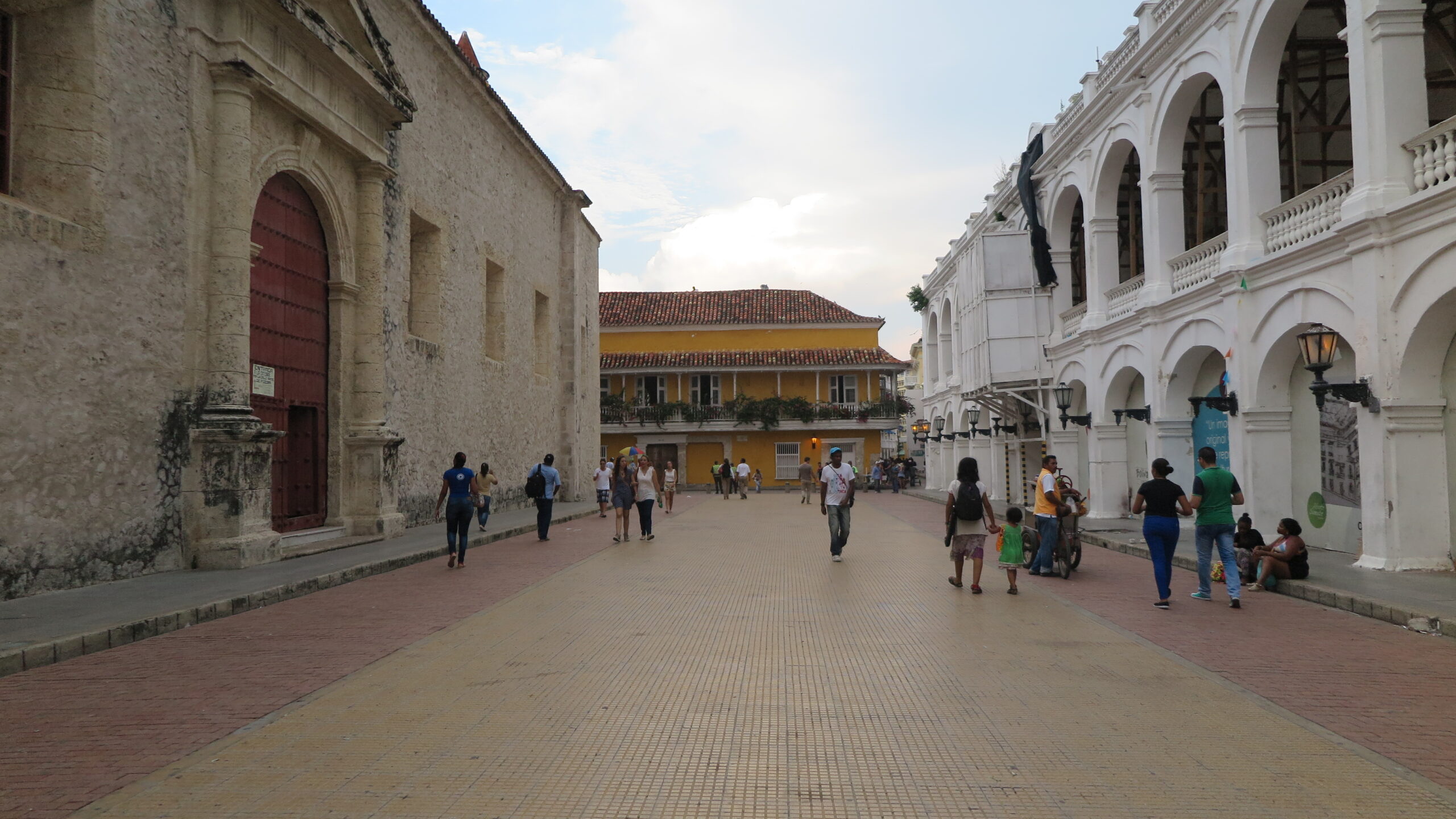 People walking through a wide, open plaza in Cartagena flanked by historic stone and white arched buildings.
