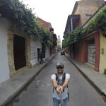 A man taking a selfie in a narrow street framed by colorful houses, with a cathedral tower visible in the distance.