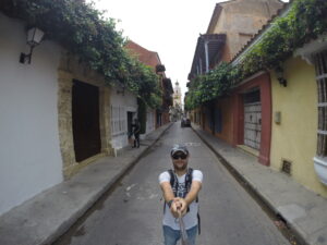 A man taking a selfie in a narrow street framed by colorful houses, with a cathedral tower visible in the distance.
