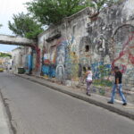A weathered wall covered in expressive graffiti and murals with people walking past in Cartagena.