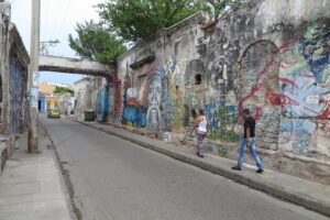 A weathered wall covered in expressive graffiti and murals with people walking past in Cartagena.