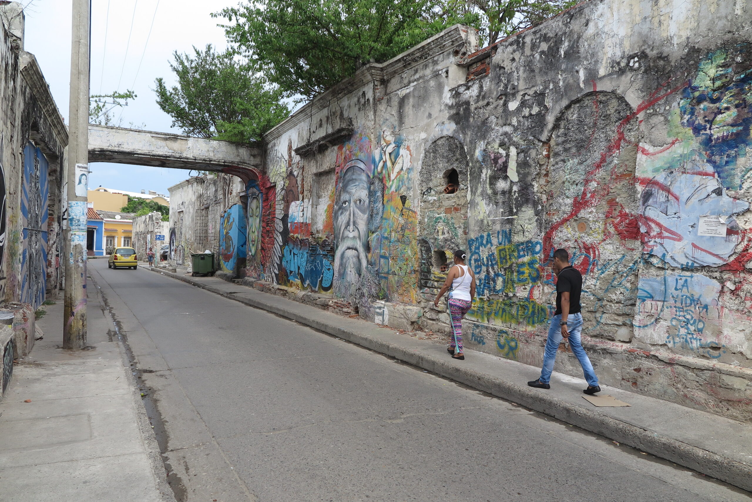 A weathered wall covered in expressive graffiti and murals with people walking past in Cartagena.