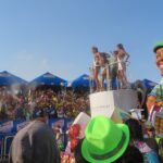 A colorful parade float in Cartagena, Colombia, featuring beauty pageant contestants waving from a platform and a giant, smiling cartoonish figure of a man in a green cap.