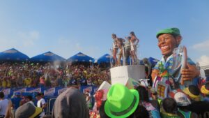 A colorful parade float in Cartagena, Colombia, featuring beauty pageant contestants waving from a platform and a giant, smiling cartoonish figure of a man in a green cap.