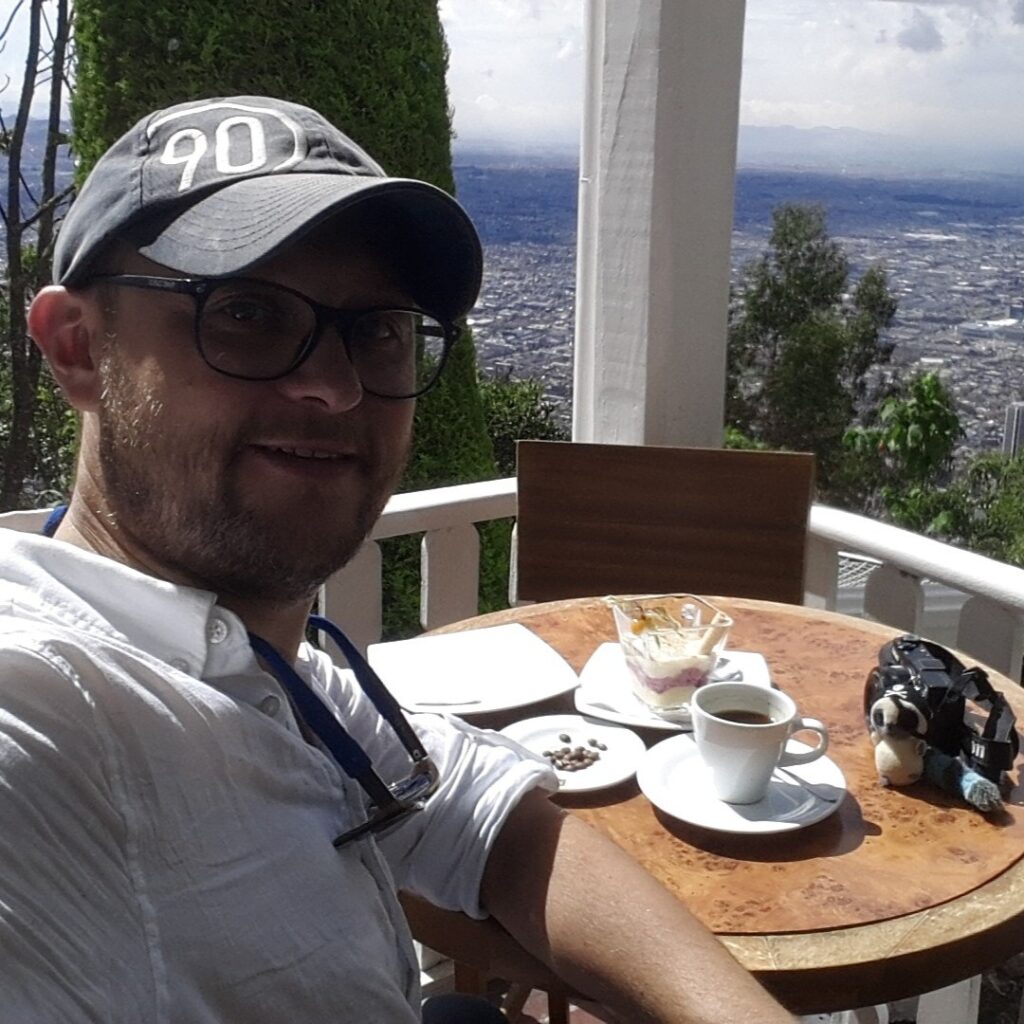 A man wearing a grey cap and glasses sitting at a cafe table on Mount Monserrate, Bogota, with a cup of coffee and a dessert. The vast city of Bogota is visible in the background under a cloudy sky.