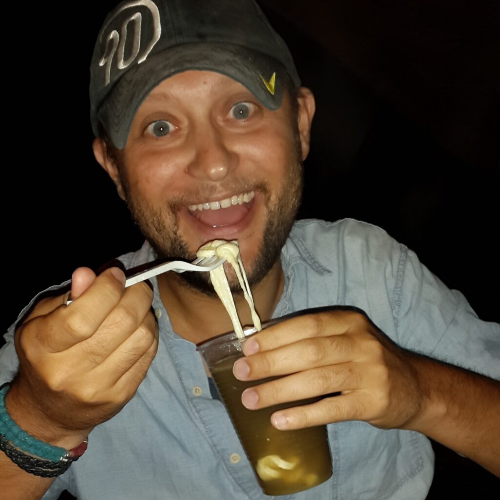 Medellin Aguapanela con queso A man with a joyful expression and wide eyes holding a plastic fork with melted, stringy cheese over a plastic cup filled with Aguapanela. He is wearing a grey cap and a light blue shirt.