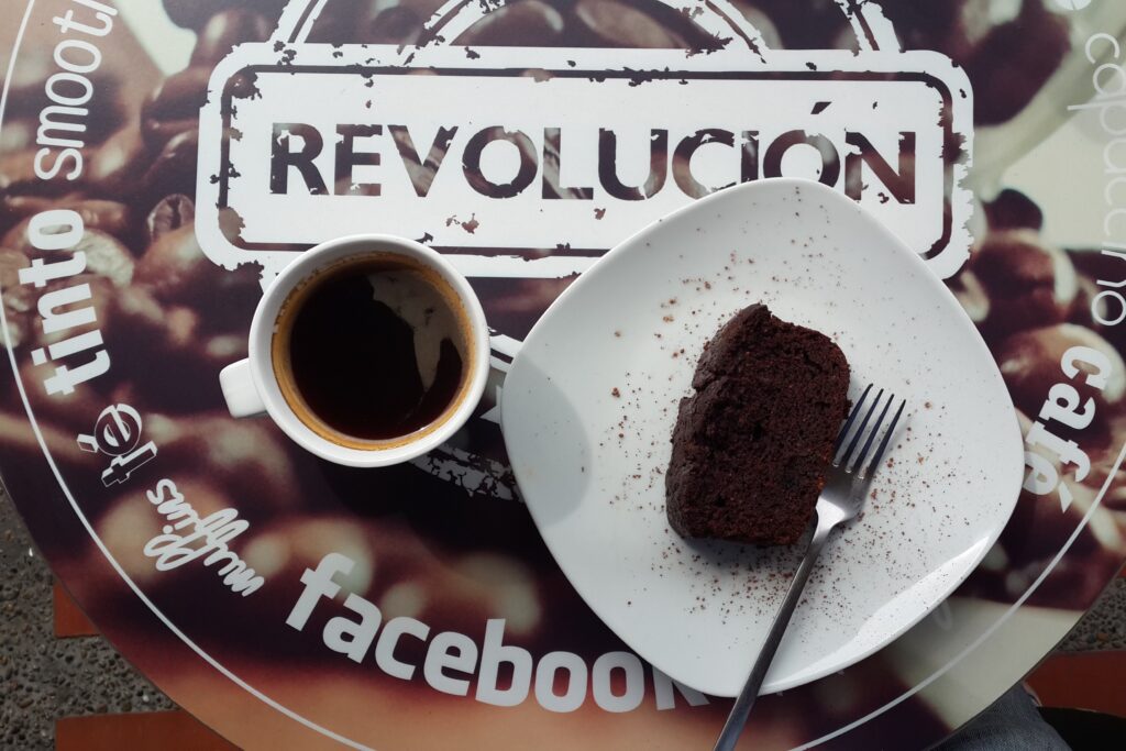Medellin Cafe Revolucion A top-down view of a white cup of black coffee and a square plate with a slice of chocolate cake on a table featuring the "Café Revolución" logo.