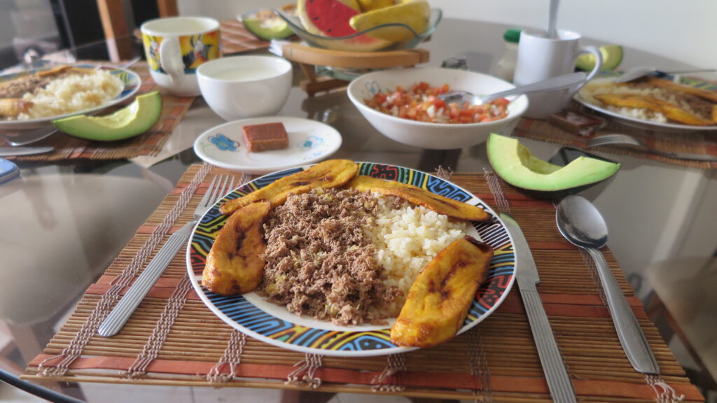 Medellin Carne Molida A colorful plate of Carne Molida (ground meat) served with white rice and fried plantains on a glass table. The table is set with other Colombian sides like avocado, salsa, and fruit.