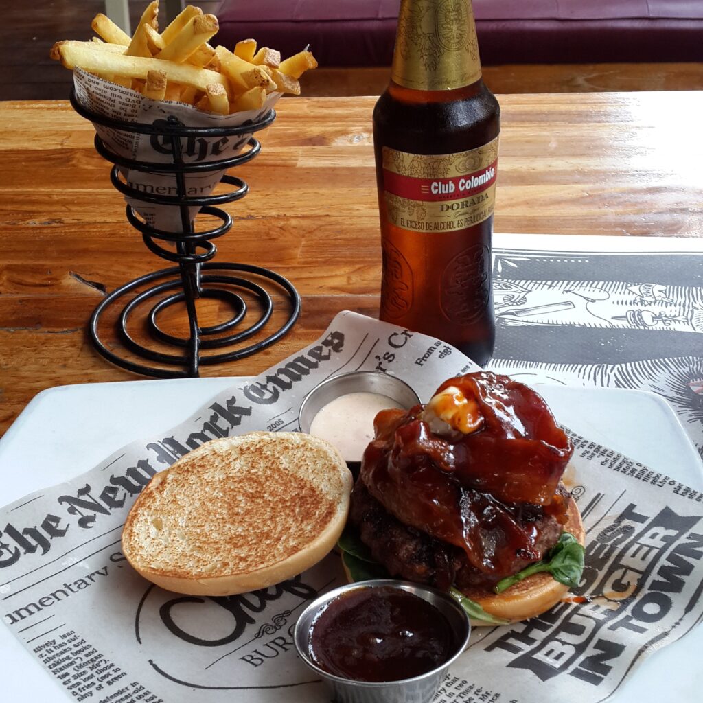 Medellin Chef Burger A gourmet bacon cheeseburger served open-faced on newspaper-print parchment paper, accompanied by a cone of french fries and a bottle of Club Colombia Dorada beer on a wooden table.