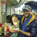 A man in a blue puffer jacket and grey cap smiling at a street food stall in Medellin while squeezing a large amount of yellow sauce onto a loaded hot dog. A menu and various sauce bottles are visible in the background.
