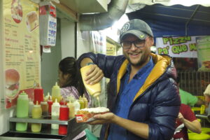A man in a blue puffer jacket and grey cap smiling at a street food stall in Medellin while squeezing a large amount of yellow sauce onto a loaded hot dog. A menu and various sauce bottles are visible in the background.