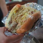 A close-up of a loaded Colombian-style street burger held in a hand. The burger is wrapped in foil and overflowing with melted cheese, sauces, and a thick layer of crispy shoestring potatoes (papitas fritas).
