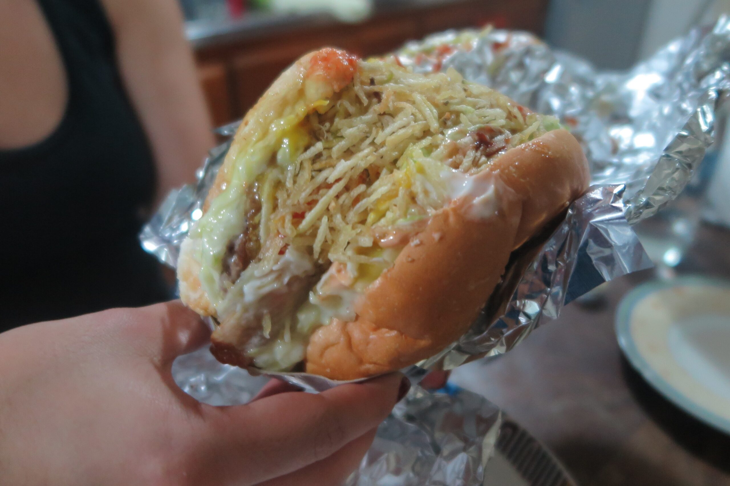 A close-up of a loaded Colombian-style street burger held in a hand. The burger is wrapped in foil and overflowing with melted cheese, sauces, and a thick layer of crispy shoestring potatoes (papitas fritas).