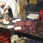 A street food vendor in Medellin grilling thin steaks and white arepas over hot charcoal on a large outdoor metal grate. The vendor is using tongs to flip an arepa.