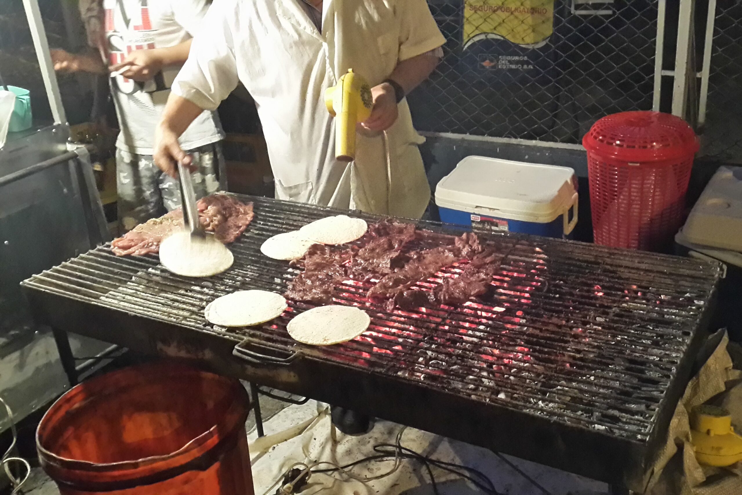 A street food vendor in Medellin grilling thin steaks and white arepas over hot charcoal on a large outdoor metal grate. The vendor is using tongs to flip an arepa.