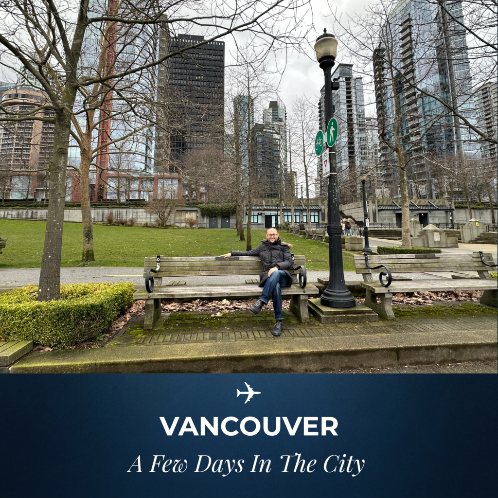 A man in a jacket sitting on a wooden bench at Harbour Green Park, Vancouver, with downtown skyscrapers and bare trees in the background on a cloudy day.
