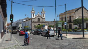 A bustling cobblestone street scene in the historic center of Ibarra, Ecuador, with the two-towered San Agustín Church in the background under a clear blue sky. Pedestrians cross the street, and cars are driving past.
