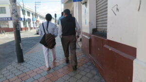 A view from behind of three people with indigenous-style braided hair, wearing traditional clothing (white pants, dark vests, shirts), walking down a paved sidewalk in a city in Ecuador. Storefronts with shutters are on the right.