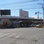 A wide-angle street view of the Mercado Amazonas building in Ibarra, Ecuador, showing a cobblestone street, local traffic with yellow taxis, and outdoor market stalls under a clear blue sky.