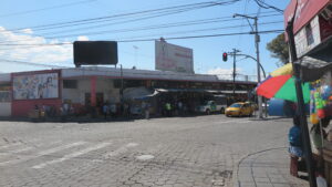 A wide-angle street view of the Mercado Amazonas building in Ibarra, Ecuador, showing a cobblestone street, local traffic with yellow taxis, and outdoor market stalls under a clear blue sky.