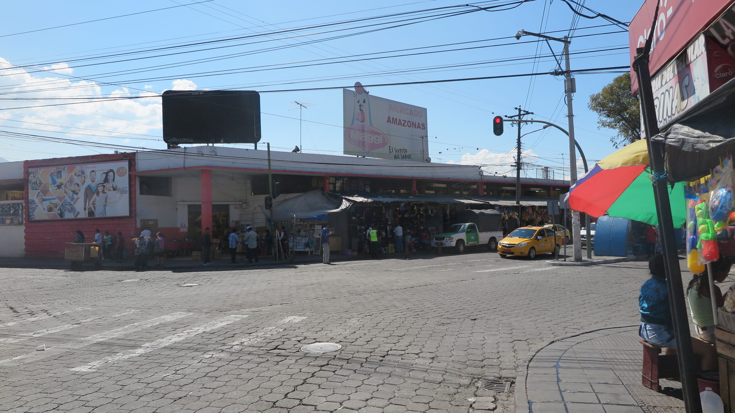 A wide-angle street view of the Mercado Amazonas building in Ibarra, Ecuador, showing a cobblestone street, local traffic with yellow taxis, and outdoor market stalls under a clear blue sky.