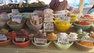 A vibrant display of various bulk spices, herbs, and roots at a market stall in Mercado Amazonas, Ibarra, Ecuador, with hand-written labels such as Ajo Molido, Linaza, and Jengibre.