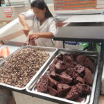 A local woman at a food stall in Mercado Amazonas, Ibarra, Ecuador, preparing traditional ingredients including chopped offal and dark blood cubes in large metal trays.