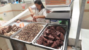A local woman at a food stall in Mercado Amazonas, Ibarra, Ecuador, preparing traditional ingredients including chopped offal and dark blood cubes in large metal trays.