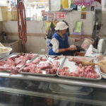 An authentic market stall inside Mercado Amazonas in Ibarra, Ecuador, where a woman in a blue vest and white hat prepares meat while an indigenous man in a fedora hat sits nearby. Various cuts of pork and lard are displayed on metal trays.