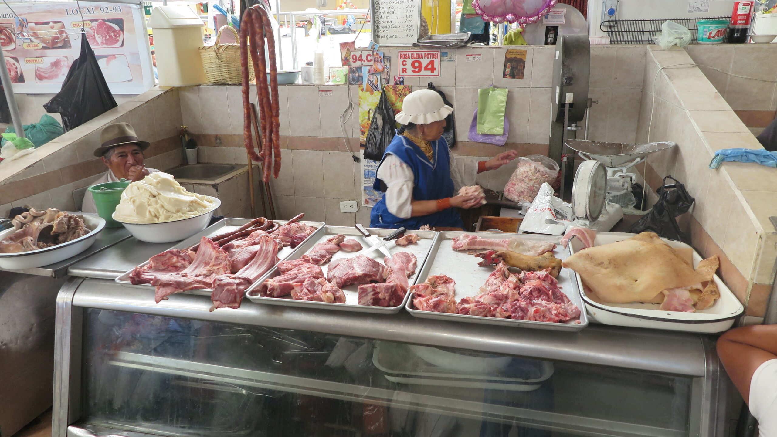 An authentic market stall inside Mercado Amazonas in Ibarra, Ecuador, where a woman in a blue vest and white hat prepares meat while an indigenous man in a fedora hat sits nearby. Various cuts of pork and lard are displayed on metal trays.