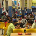 A wide-angle interior view of the bustling open-air food court within Mercado Amazonas in Ibarra, Ecuador. Numerous locals are seated at simple wooden tables and bright yellow plastic chairs, enjoying traditional meals. The space features large pillars with colorful advertisements and various food stalls.