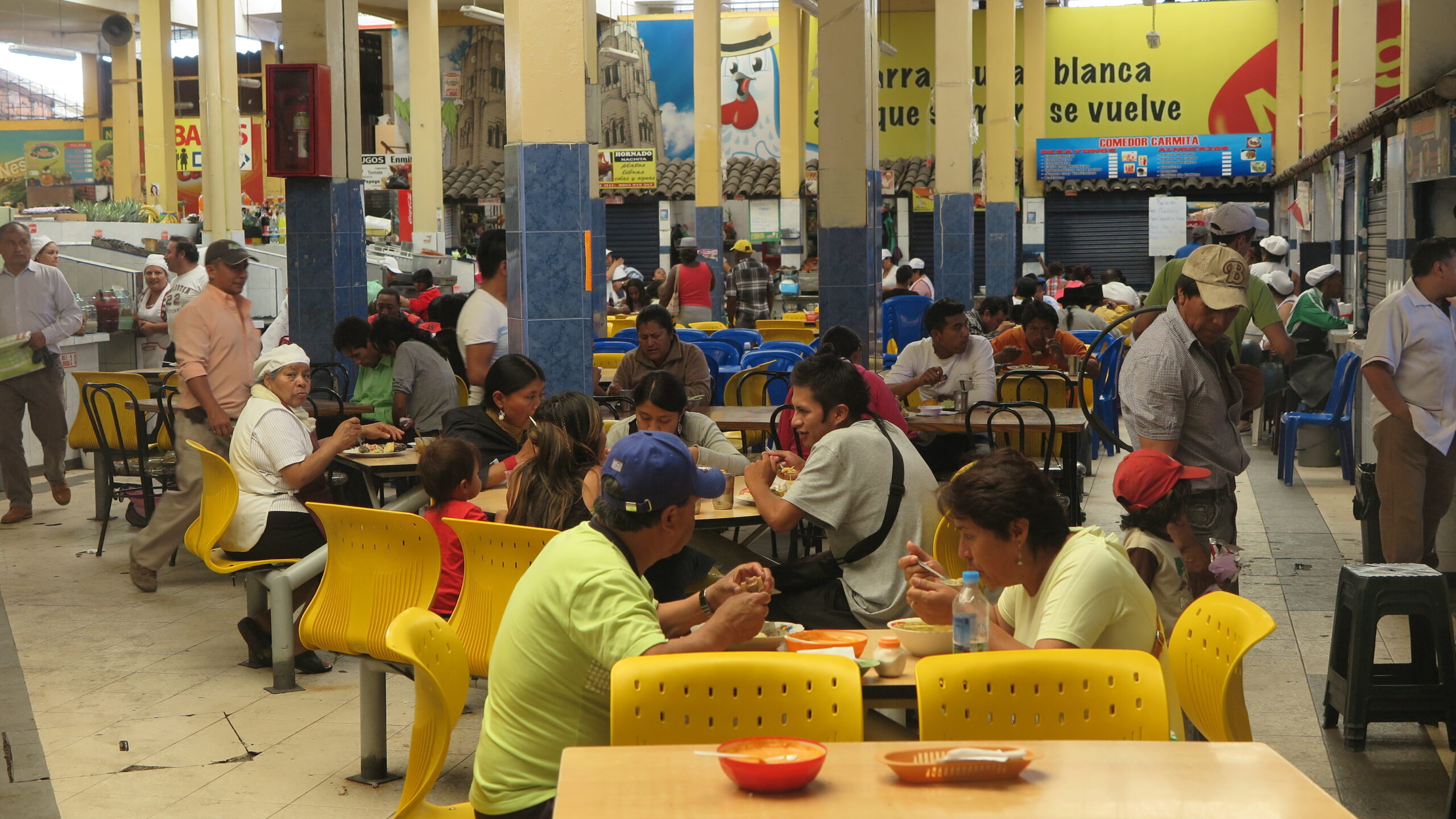 A wide-angle interior view of the bustling open-air food court within Mercado Amazonas in Ibarra, Ecuador. Numerous locals are seated at simple wooden tables and bright yellow plastic chairs, enjoying traditional meals. The space features large pillars with colorful advertisements and various food stalls.