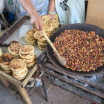 A high-angle shot of a vendor at Mercado Amazonas in Ibarra, Ecuador, roasting broad beans in a large clay pot over a charcoal brazier, with stacks of toasted corn tortillas on a wooden tray nearby.