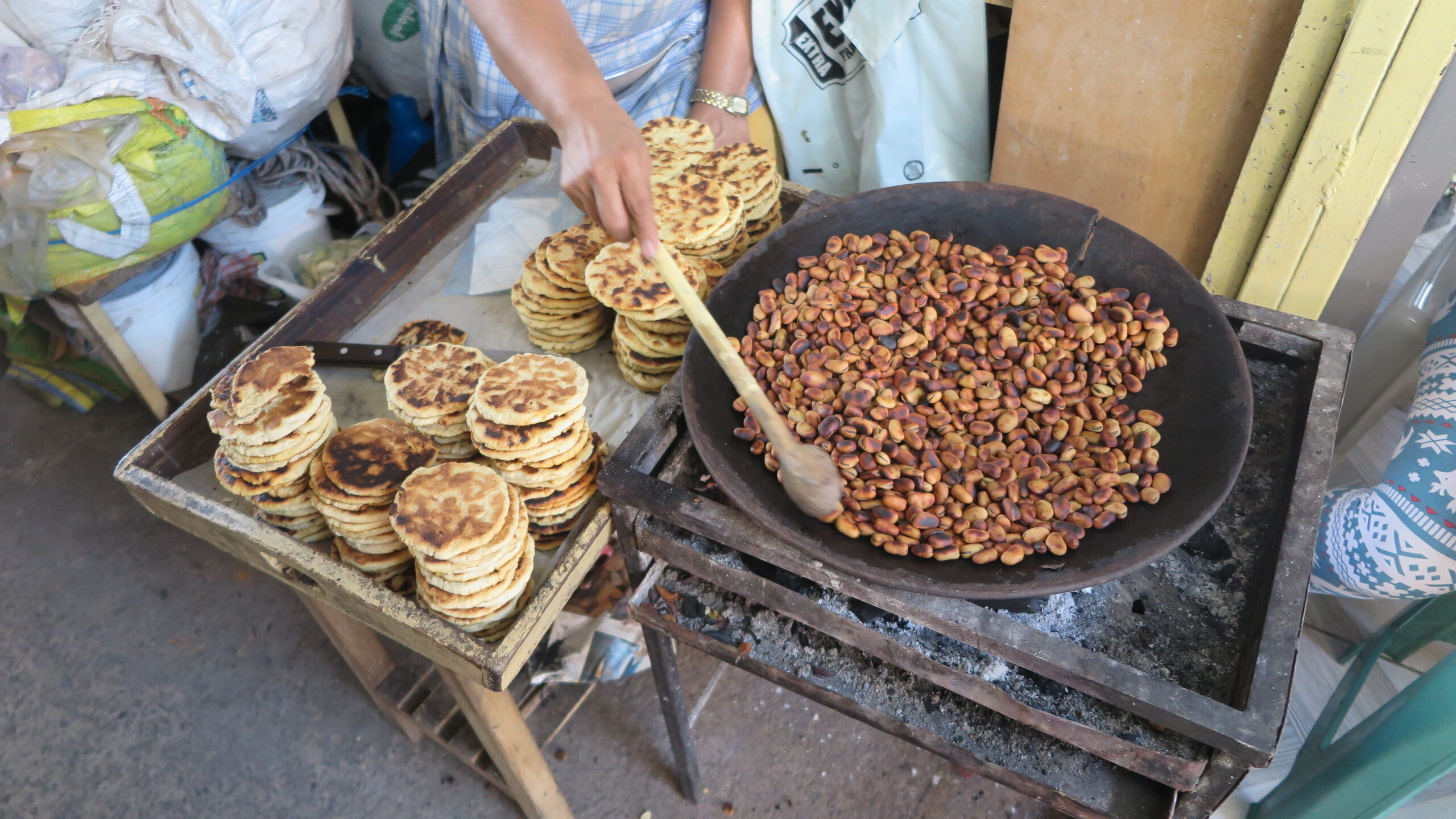 A high-angle shot of a vendor at Mercado Amazonas in Ibarra, Ecuador, roasting broad beans in a large clay pot over a charcoal brazier, with stacks of toasted corn tortillas on a wooden tray nearby.