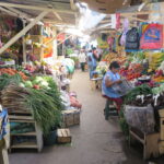 A vibrant and narrow aisle inside Mercado Amazonas in Ibarra, Ecuador, lined with overflowing stalls of fresh tropical fruits, vegetables, and tubers. A local vendor in a blue apron is seen sitting and reading a newspaper amidst the produce.