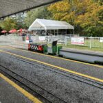 A miniature red and black locomotive waiting at the Burnaby Central Railway station platform, with autumn trees and a white tent in the background.