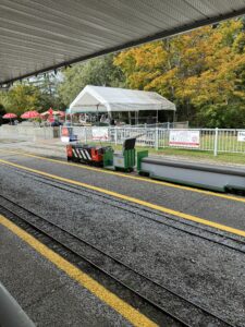 A miniature red and black locomotive waiting at the Burnaby Central Railway station platform, with autumn trees and a white tent in the background.