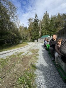 People sitting on a miniature train traveling through a forested area at Burnaby Central Railway, Vancouver, under a blue sky with white clouds.