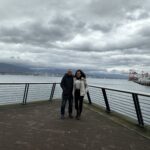 A couple posing on the Canada Place pier in Vancouver, with the ocean and harbor cranes in the background.