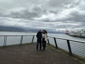 A couple posing on the Canada Place pier in Vancouver, with the ocean and harbor cranes in the background.